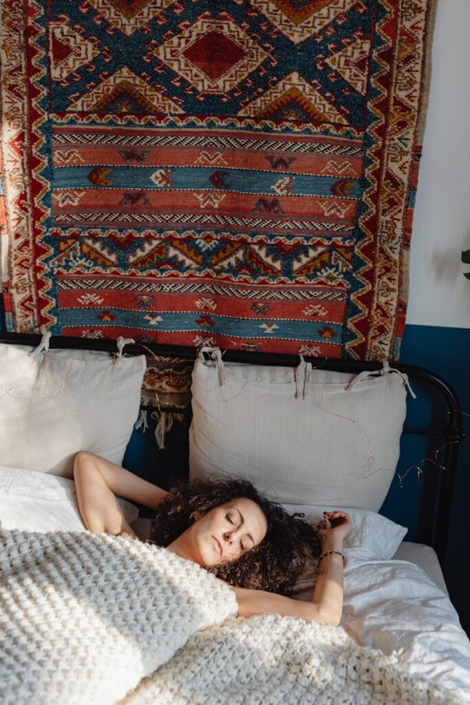 A peaceful woman rests in a cozy bedroom with a decorative tapestry on the wall, sunlight streaming in.